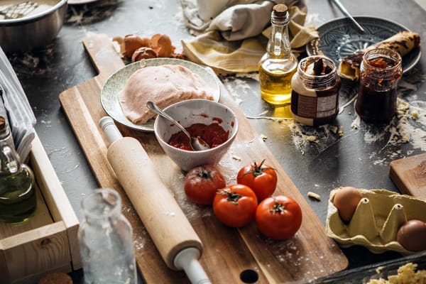 A counter that has grown quite dirty from chaotic cooking, containing ingredients in various stages of prperation. Jame in an jar, oil in a bottle, some tomatoes, tomato paste etc